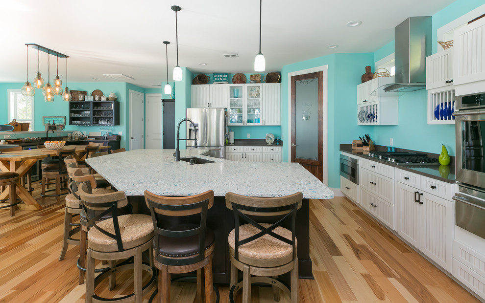Large beach style l-shaped open plan kitchen in Charleston with a single-bowl sink, louvered cabinets, white cabinets, recycled glass benchtops, blue splashback, stainless steel appliances, medium hardwood floors, with island and beige floor.