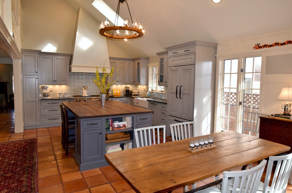 This is an example of a transitional l-shaped open plan kitchen in Denver with a farmhouse sink, recessed-panel cabinets, grey cabinets, wood benchtops, grey splashback, glass tile splashback, panelled appliances, terra-cotta floors and with island.