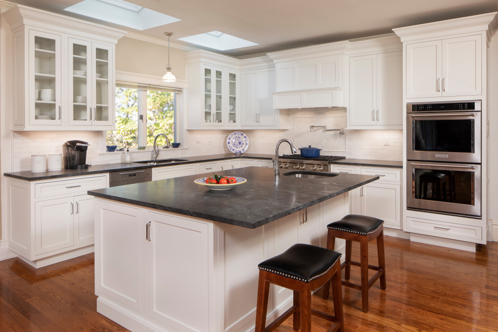 Inspiration for a large traditional l-shaped eat-in kitchen in New York with an undermount sink, shaker cabinets, white cabinets, granite benchtops, white splashback, subway tile splashback, stainless steel appliances, medium hardwood floors, with island, beige floor, black benchtop and exposed beam.
