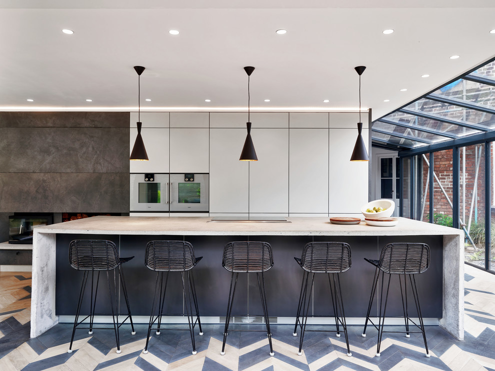 This is an example of a contemporary galley open plan kitchen in Devon with flat-panel cabinets, grey cabinets, concrete benchtops, stainless steel appliances, light hardwood floors, with island, multi-coloured floor and grey benchtop.