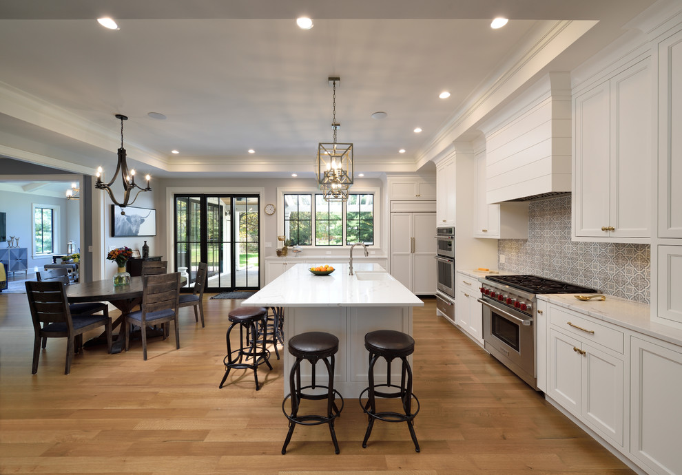 Inspiration for a large country l-shaped separate kitchen in Chicago with a farmhouse sink, recessed-panel cabinets, white cabinets, quartz benchtops, brown splashback, mosaic tile splashback, panelled appliances, light hardwood floors, with island, brown floor and white benchtop.