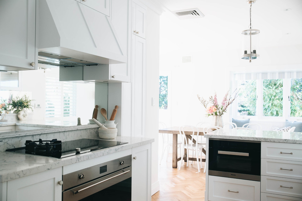 Design ideas for a mid-sized traditional l-shaped eat-in kitchen in Sydney with an undermount sink, recessed-panel cabinets, white cabinets, quartz benchtops, black appliances, medium hardwood floors, white benchtop and mirror splashback.
