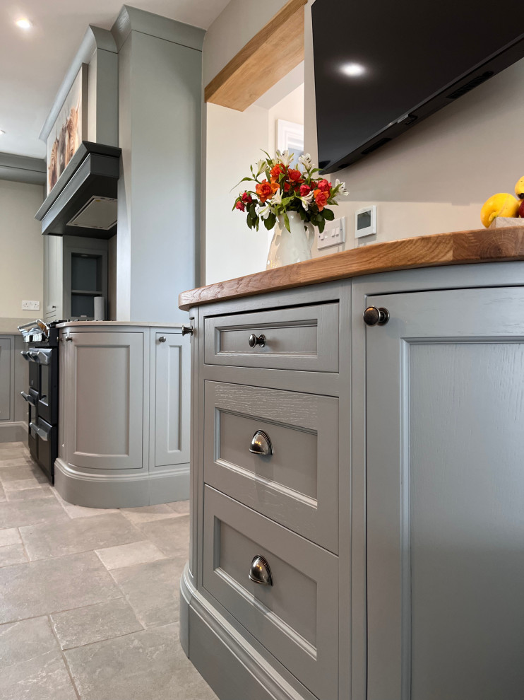 This is an example of a large country u-shaped eat-in kitchen in Essex with a farmhouse sink, shaker cabinets, grey cabinets, wood benchtops, grey splashback, black appliances, with island and brown benchtop.