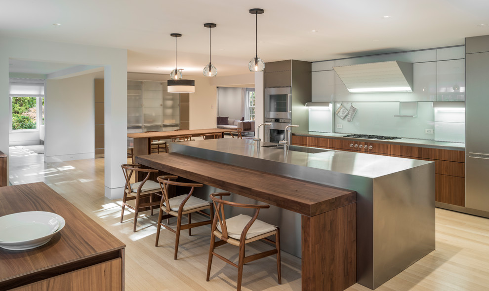Contemporary kitchen in Boston with a double-bowl sink, flat-panel cabinets, dark wood cabinets, white splashback, stainless steel appliances, light hardwood floors, with island and grey benchtop.