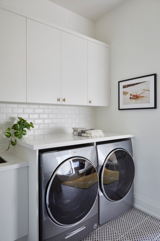 トロントにある高級な広いトランジショナルスタイルのおしゃれな家事室 (I型、アンダーカウンターシンク、フラットパネル扉のキャビネット、白いキャビネット、人工大理石カウンター、白いキッチンパネル、磁器タイルのキッチンパネル、白い壁、セラミックタイルの床、左右配置の洗濯機・乾燥機、マルチカラーの床、白いキッチンカウンター) の写真
