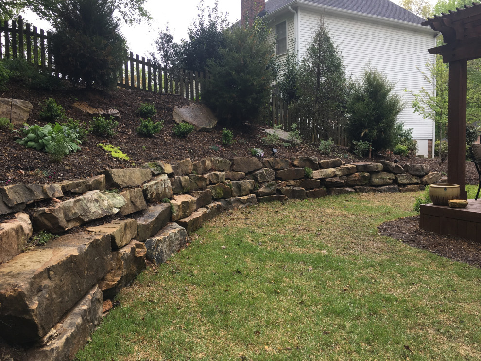 Photo of a mid-sized traditional backyard partial sun xeriscape in Other with a retaining wall, natural stone pavers and a stone fence.