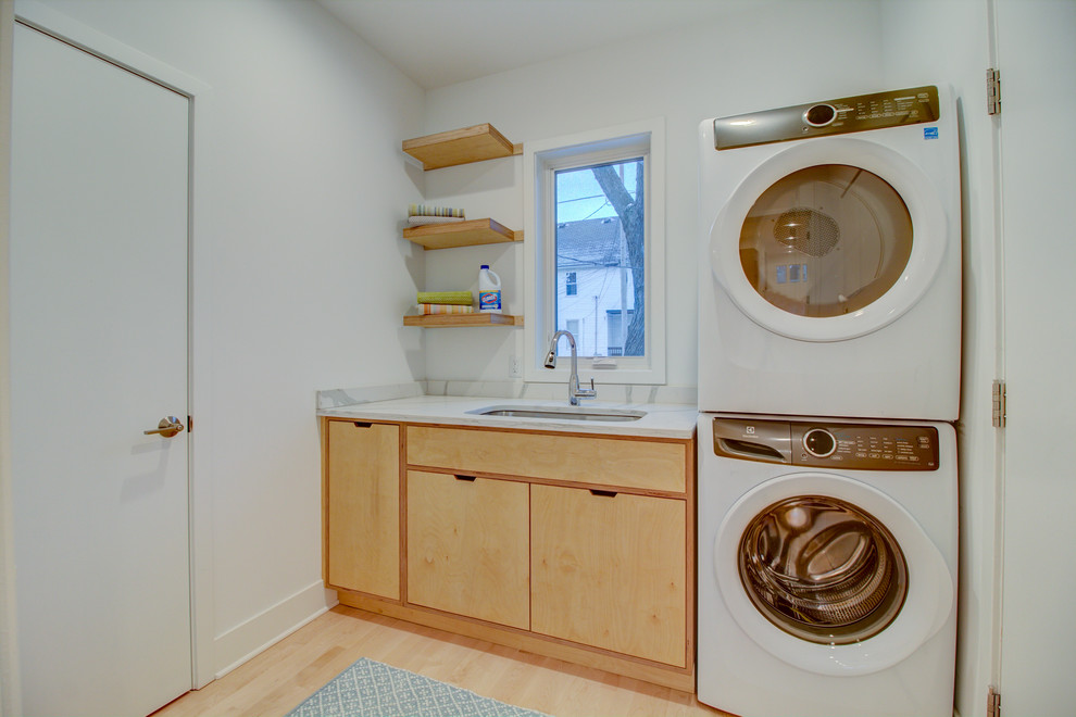 Design ideas for a small modern single-wall utility room in Milwaukee with an undermount sink, flat-panel cabinets, light wood cabinets, quartz benchtops, white walls, light hardwood floors, a stacked washer and dryer, brown floor and white benchtop.