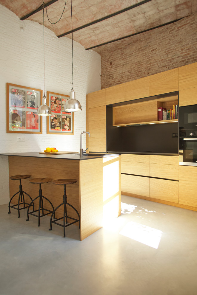 Photo of a mid-sized mediterranean galley eat-in kitchen in Barcelona with medium wood cabinets, black splashback, black appliances, concrete floors, with island, grey floor, black benchtop, an undermount sink and flat-panel cabinets.