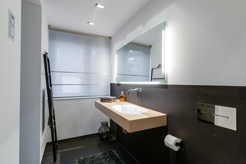 This is an example of a small modern 3/4 bathroom in Cologne with a curbless shower, a wall-mount toilet, wood benchtops, an open shower, beige benchtops, beaded inset cabinets, light wood cabinets, black tile, matchstick tile, white walls, an integrated sink, black floor, a niche, a single vanity, a floating vanity and recessed.