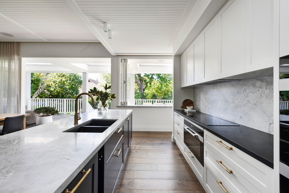Mid-sized beach style open plan kitchen in Sydney with a drop-in sink, shaker cabinets, white cabinets, marble benchtops, grey splashback, marble splashback, black appliances, medium hardwood floors, with island, brown floor, grey benchtop and timber.