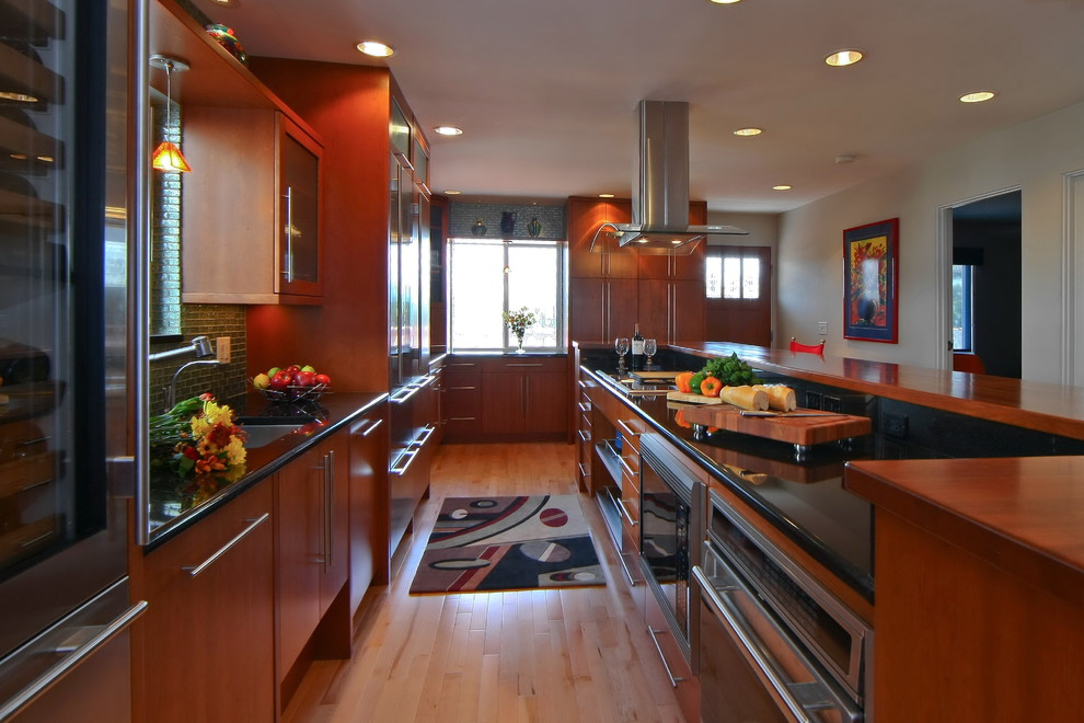 Example of a large transitional l-shaped light wood floor eat-in kitchen design with an undermount sink, flat-panel cabinets, medium tone wood cabinets, wood countertops, green backsplash, glass tile backsplash, stainless steel appliances and an island