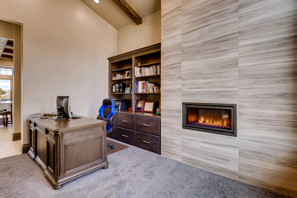Photo of a mid-sized transitional home office in Denver with a library, beige walls, carpet, a ribbon fireplace, a tile fireplace surround, a freestanding desk and grey floor.