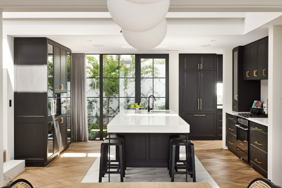 This is an example of a transitional u-shaped eat-in kitchen with black cabinets, light hardwood floors, white benchtop, an undermount sink, shaker cabinets, panelled appliances, with island and brown floor.