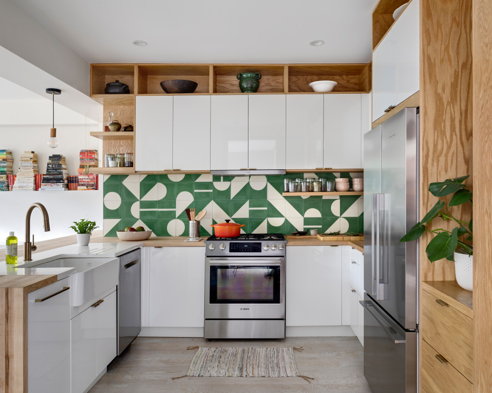 This is an example of a contemporary u-shaped kitchen in New York with a farmhouse sink, flat-panel cabinets, white cabinets, wood benchtops, green splashback, cement tile splashback, stainless steel appliances, light hardwood floors and no island.