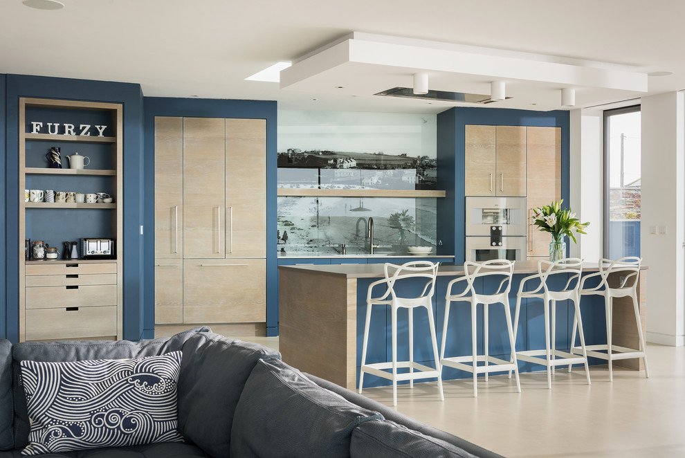 Large contemporary single-wall open plan kitchen in Cornwall with a drop-in sink, flat-panel cabinets, medium wood cabinets, quartzite benchtops, multi-coloured splashback, glass sheet splashback, panelled appliances, concrete floors, with island, beige floor and beige benchtop.