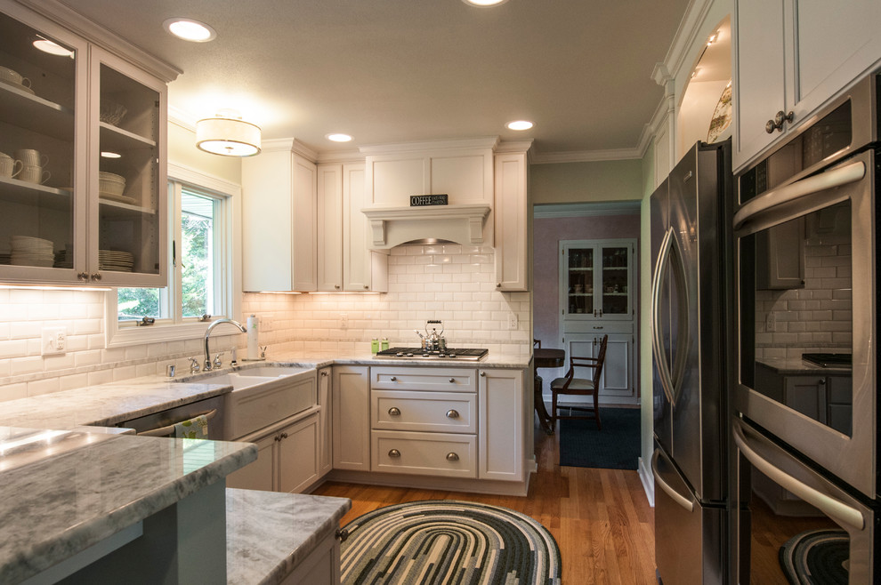 Inspiration for a small transitional u-shaped medium tone wood floor enclosed kitchen remodel in Milwaukee with a farmhouse sink, shaker cabinets, white cabinets, granite countertops, white backsplash, subway tile backsplash, stainless steel appliances and a peninsula