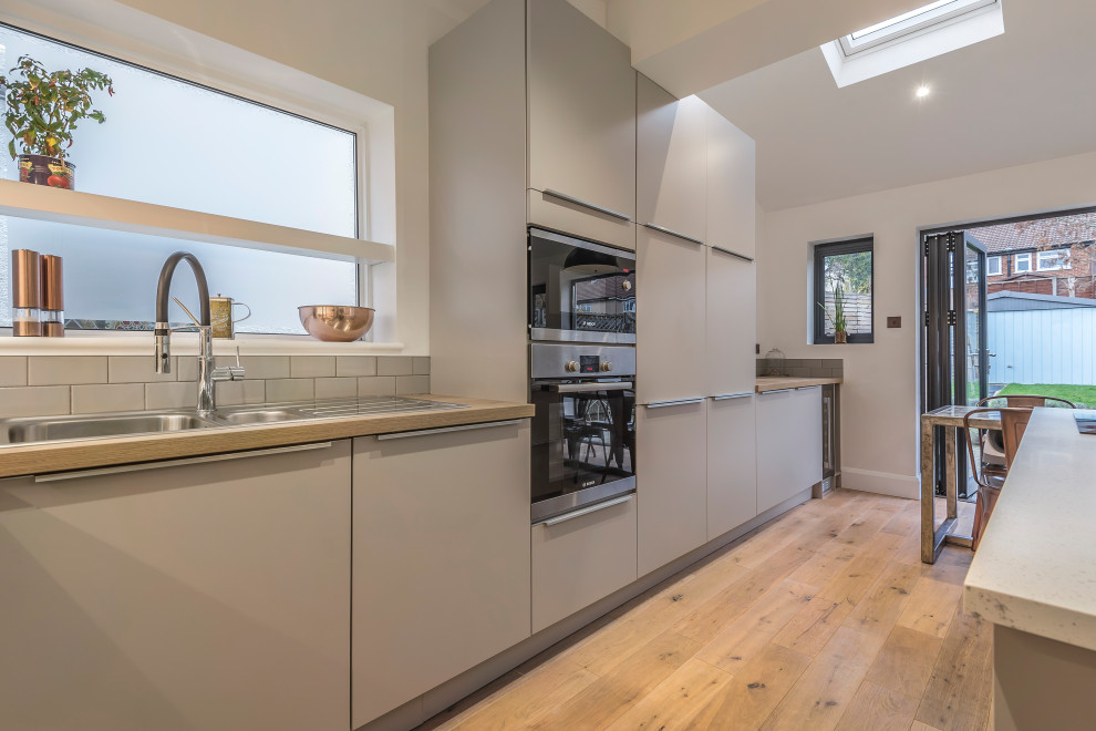 Photo of a large contemporary grey and white single-wall kitchen/diner in Kent with a built-in sink, flat-panel cabinets, grey cabinets, composite countertops, grey splashback, ceramic splashback, integrated appliances, medium hardwood flooring, an island, brown floors, white worktops, a vaulted ceiling and feature lighting.