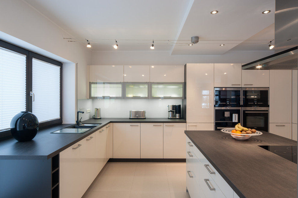 Inspiration for a large contemporary u-shaped open plan kitchen in Dresden with a double-bowl sink, flat-panel cabinets, white cabinets, glass sheet splashback, stainless steel appliances, ceramic floors, a peninsula and white splashback.
