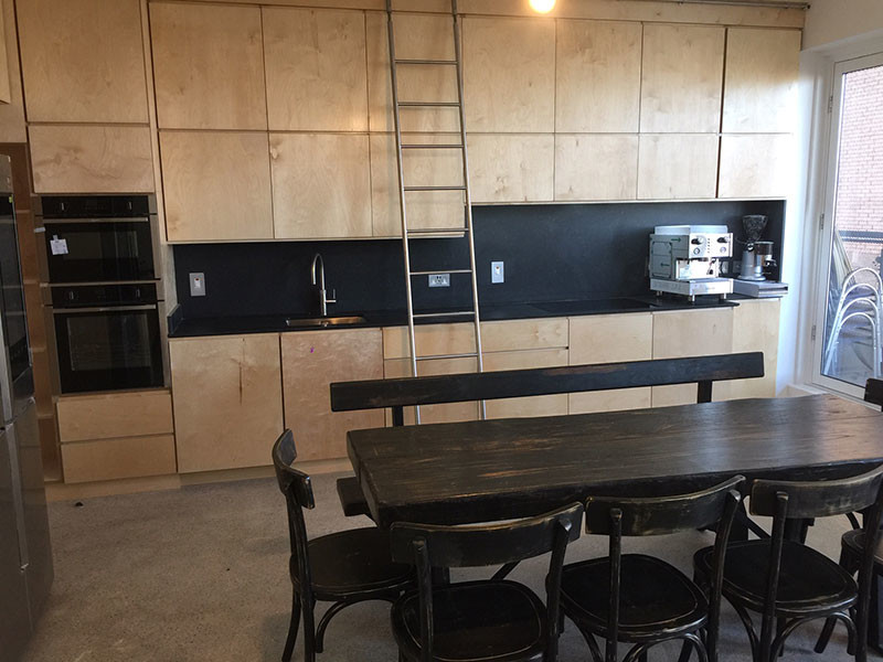 Photo of a mid-sized eclectic l-shaped eat-in kitchen in Dublin with an integrated sink, flat-panel cabinets, light wood cabinets, quartzite benchtops, black splashback, slate splashback, black appliances, concrete floors, no island, grey floor and black benchtop.