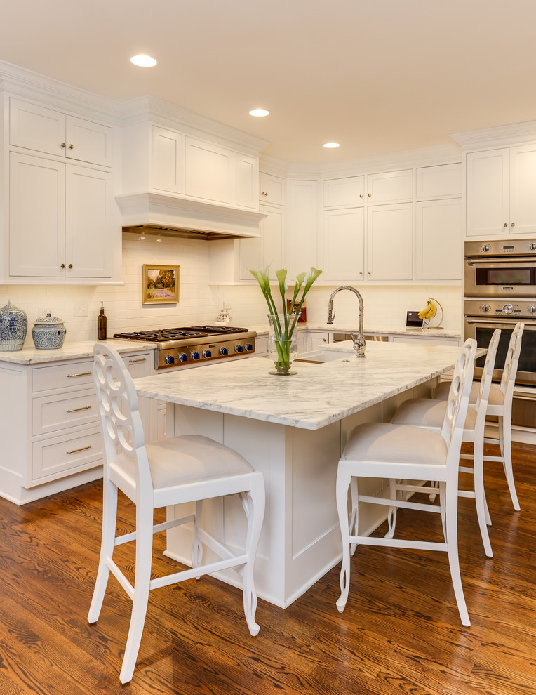 Mid-sized traditional l-shaped kitchen in Raleigh with a farmhouse sink, white cabinets, marble benchtops, white splashback, ceramic splashback, stainless steel appliances, medium hardwood floors, with island, brown floor, white benchtop and shaker cabinets.
