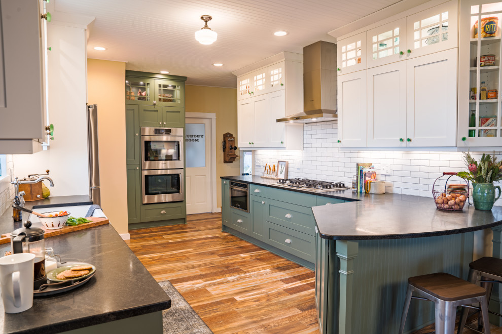 Photo of a country u-shaped kitchen/diner in Minneapolis with a belfast sink, shaker cabinets, green cabinets, white splashback, metro tiled splashback, stainless steel appliances, a breakfast bar, brown floors and black worktops.