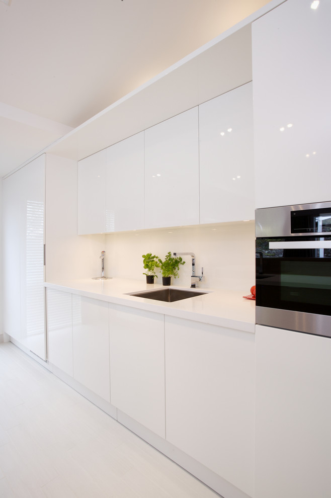 Small contemporary galley eat-in kitchen in Auckland with an undermount sink, flat-panel cabinets, white cabinets, solid surface benchtops, white splashback, glass sheet splashback, stainless steel appliances, light hardwood floors and with island.