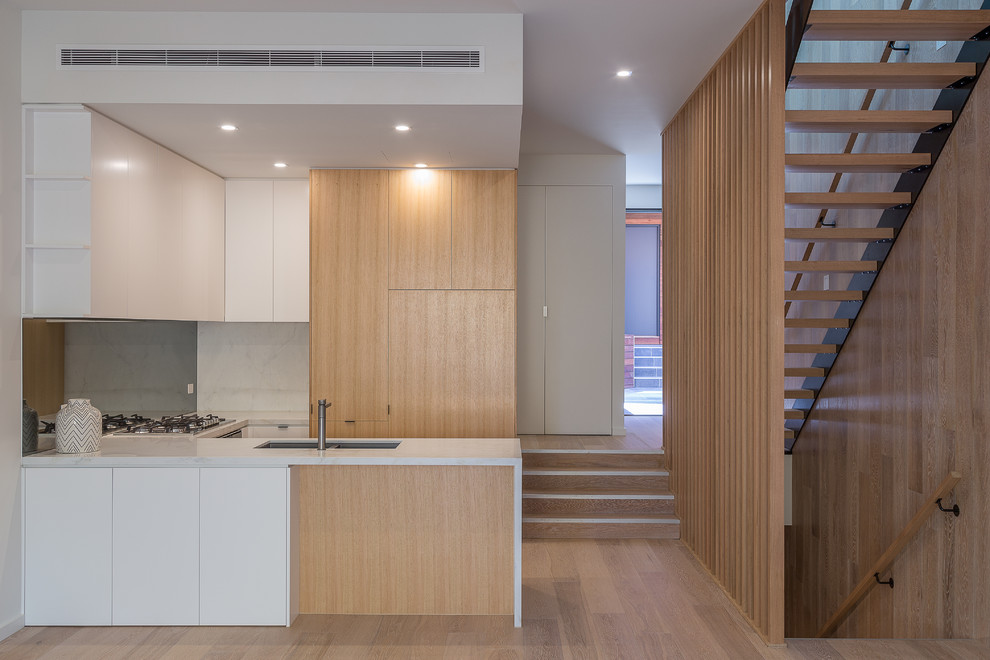 Small contemporary u-shaped open plan kitchen in Melbourne with an undermount sink, white cabinets, white splashback, stainless steel appliances, light hardwood floors, with island and brown floor.