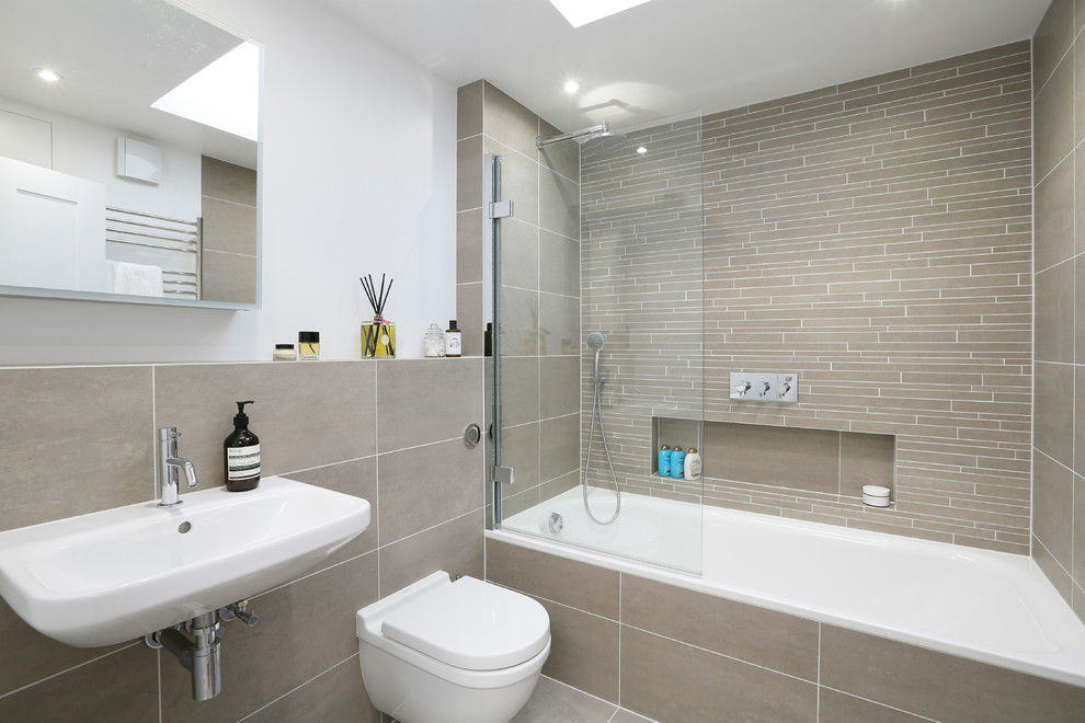 This is an example of a mid-sized contemporary kids bathroom in London with a shower/bathtub combo, a wall-mount toilet, beige tile, white walls, a wall-mount sink, a hinged shower door, a drop-in tub, ceramic floors, grey floor and a single vanity.