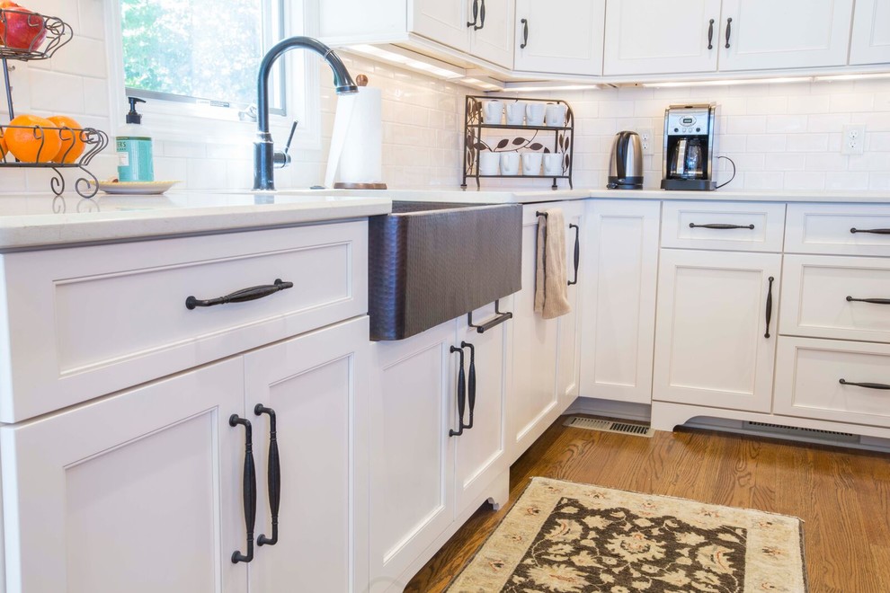 Inspiration for a large transitional l-shaped light wood floor and brown floor enclosed kitchen remodel with a farmhouse sink, shaker cabinets, white cabinets, solid surface countertops, white backsplash, porcelain backsplash, stainless steel appliances and an island