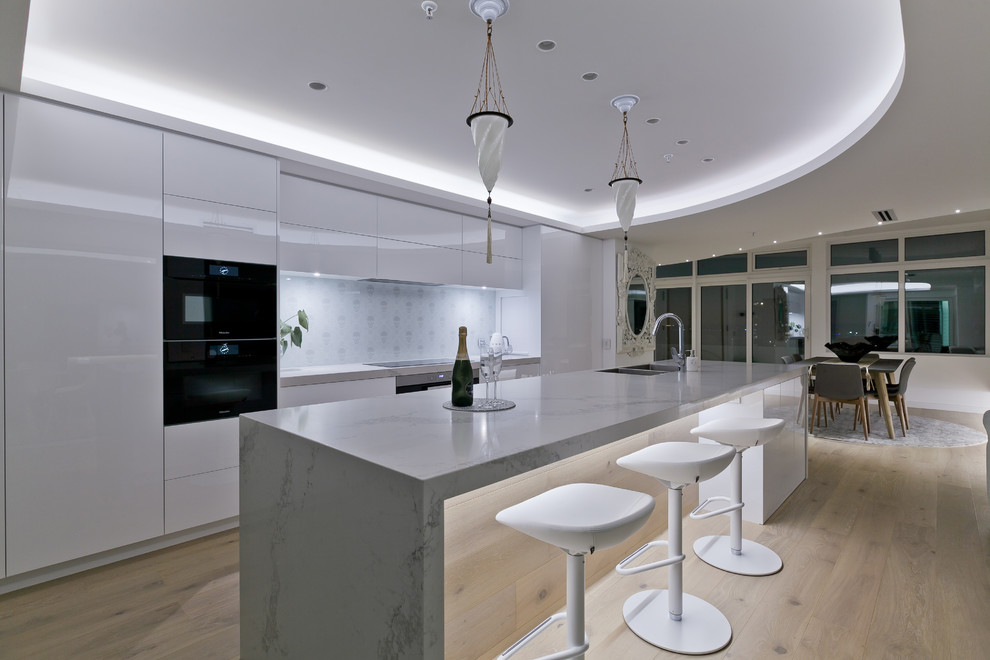 This is an example of a large contemporary galley open plan kitchen in Perth with an undermount sink, flat-panel cabinets, white cabinets, quartz benchtops, white splashback, porcelain splashback, stainless steel appliances, light hardwood floors, with island and white benchtop.