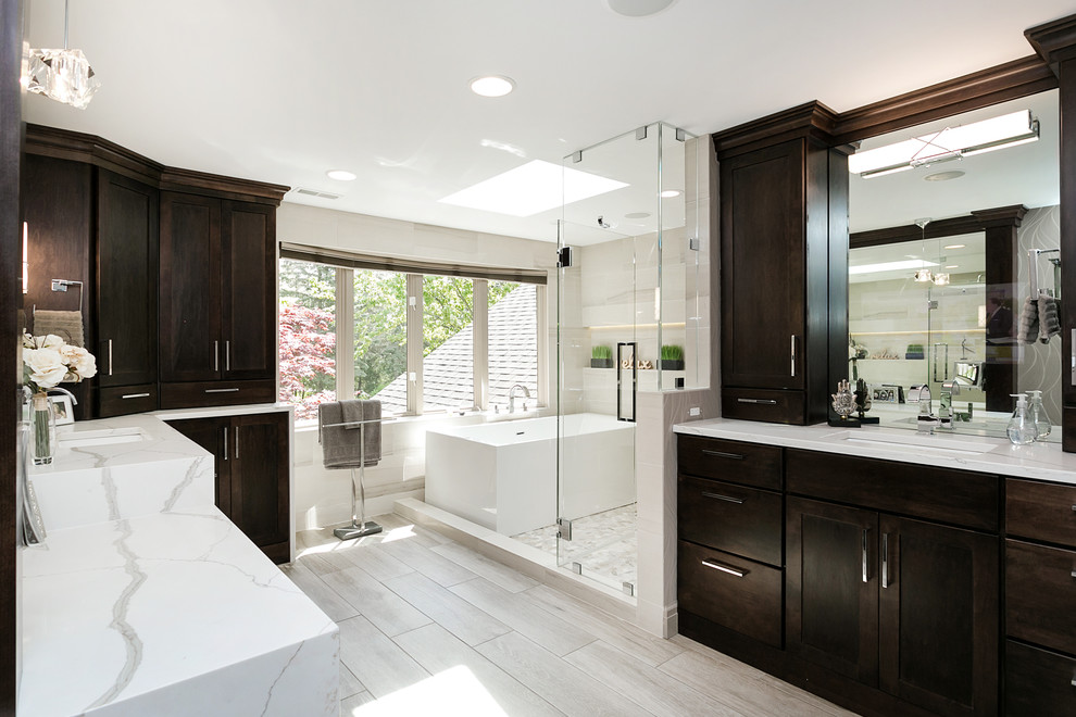 This is an example of a mid-sized transitional master bathroom in Detroit with shaker cabinets, dark wood cabinets, a freestanding tub, a shower/bathtub combo, a two-piece toilet, beige tile, porcelain tile, porcelain floors, an undermount sink, marble benchtops, brown floor, a hinged shower door and white benchtops.