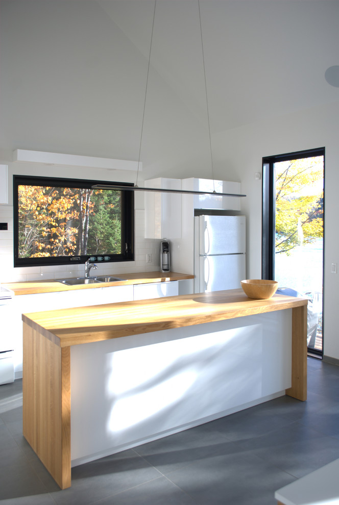 Small scandinavian galley eat-in kitchen in Montreal with an integrated sink, flat-panel cabinets, white cabinets, wood benchtops, white splashback, ceramic splashback, white appliances, ceramic floors, with island, grey floor and beige benchtop.