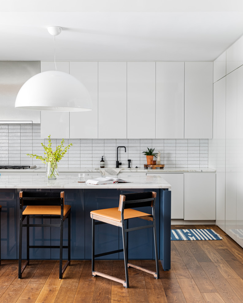 This is an example of a large contemporary single-wall open plan kitchen in New York with an undermount sink, flat-panel cabinets, white cabinets, marble benchtops, white splashback, ceramic splashback, stainless steel appliances, medium hardwood floors, with island and white benchtop.