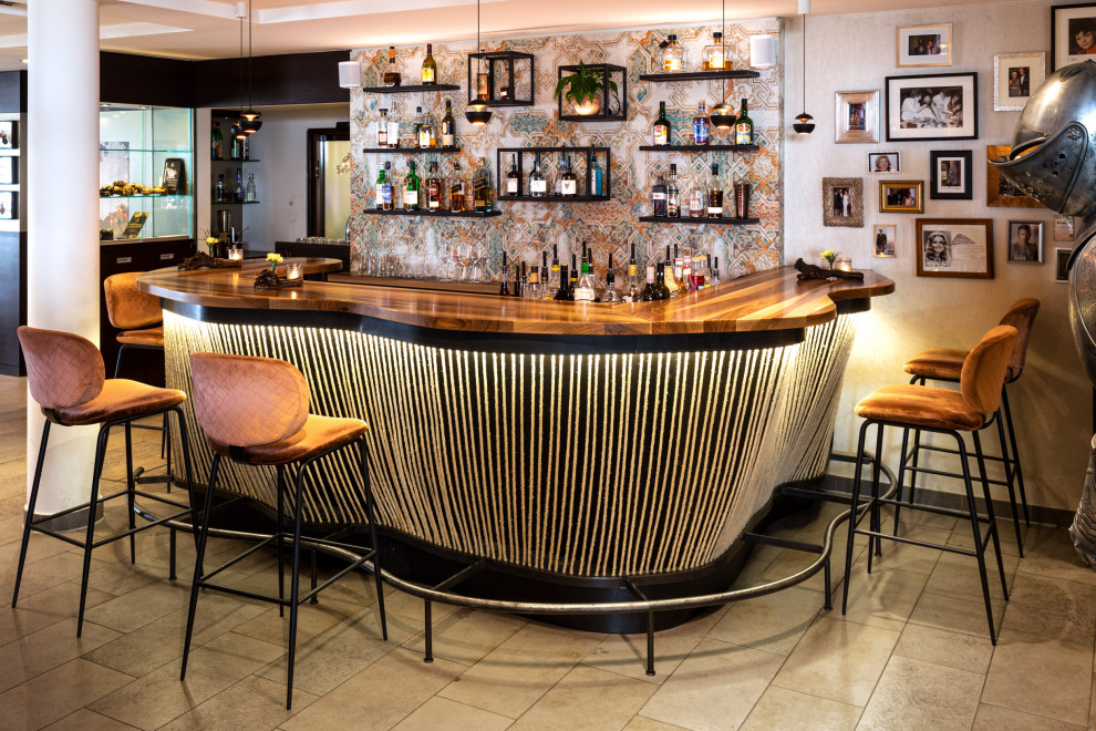 Photo of a large industrial u-shaped seated home bar in Other with an integrated sink, flat-panel cabinets, black cabinets, wood benchtops, multi-coloured splashback, subway tile splashback, travertine floors, beige floor and brown benchtop.