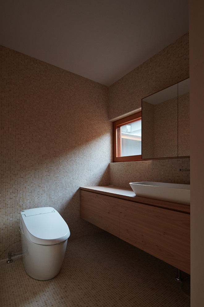 Modern powder room in Kyoto with flat-panel cabinets, light wood cabinets, a one-piece toilet, beige tile, mosaic tile, beige walls, mosaic tile floors, a vessel sink, wood benchtops and beige floor.