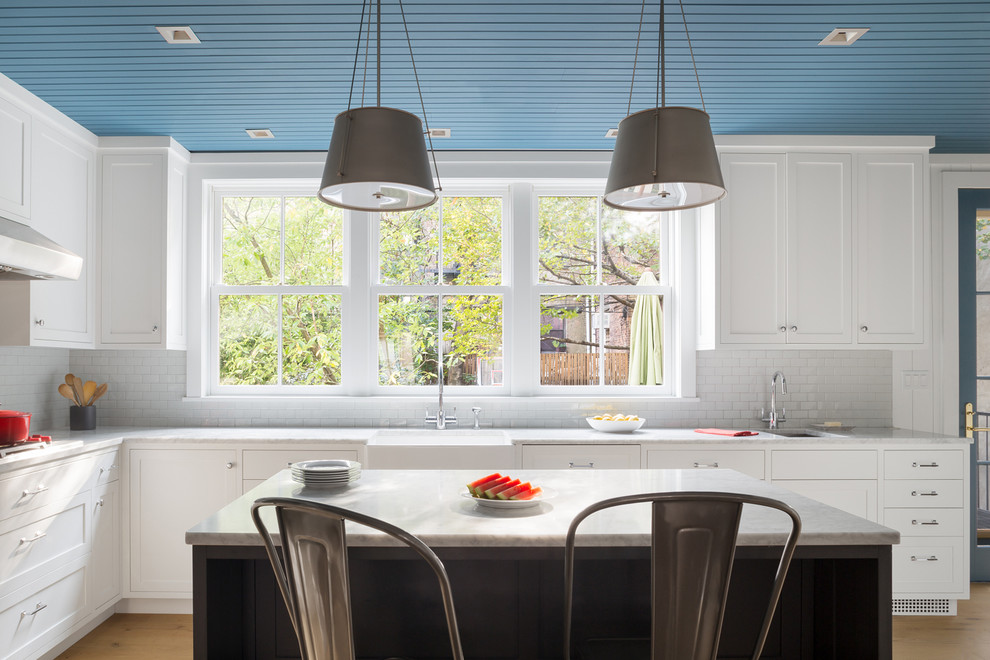 Example of a transitional l-shaped light wood floor and beige floor kitchen design in New York with a farmhouse sink, shaker cabinets, white cabinets, subway tile backsplash, an island and gray countertops