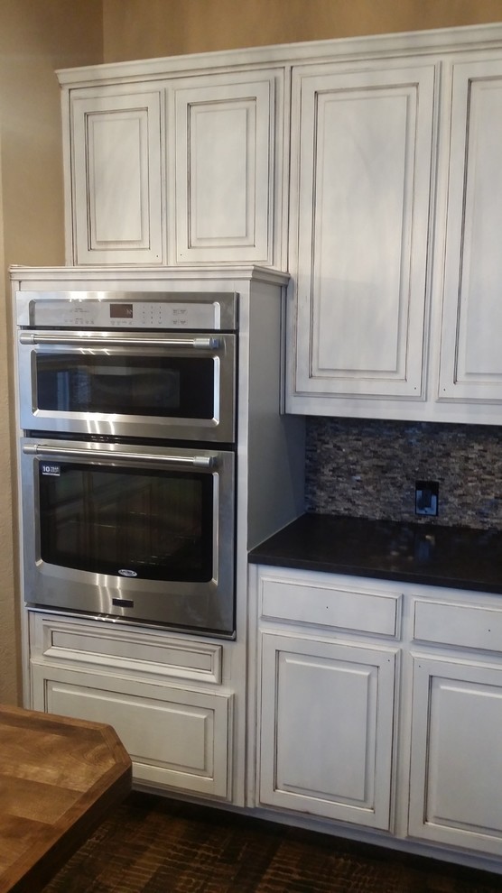 Open concept kitchen - large transitional brown floor open concept kitchen idea in Dallas with an undermount sink, raised-panel cabinets, white cabinets, granite countertops, gray backsplash, stone tile backsplash, stainless steel appliances and an island