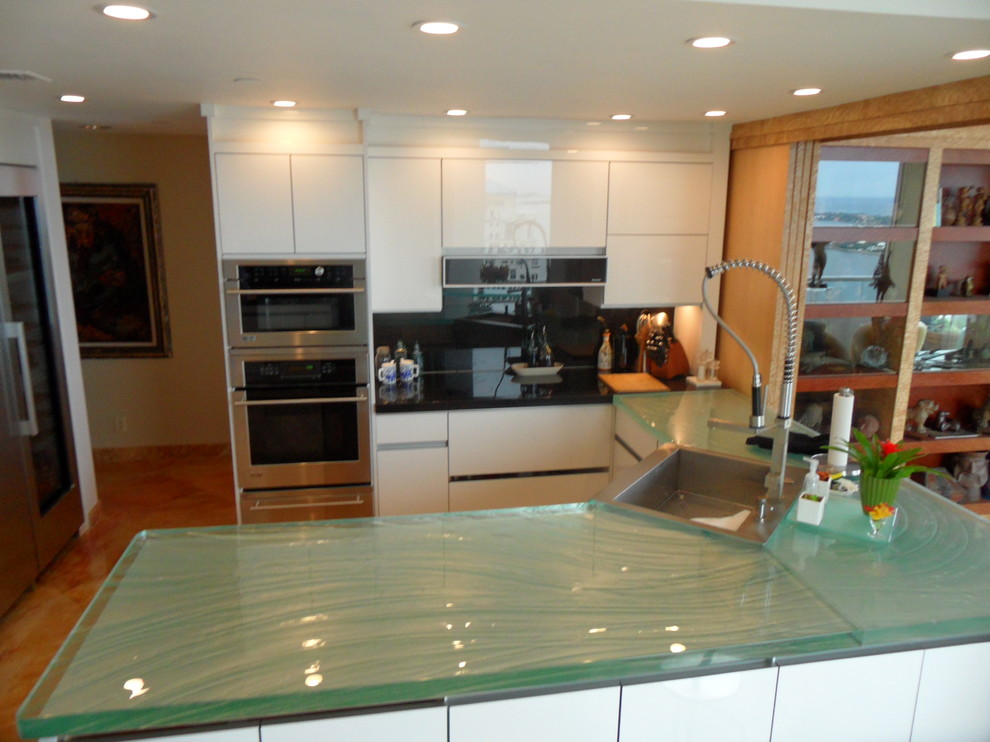 Large modern u-shaped separate kitchen in Miami with a drop-in sink, flat-panel cabinets, glass benchtops, black splashback, glass sheet splashback, stainless steel appliances, white cabinets, a peninsula and orange floor.