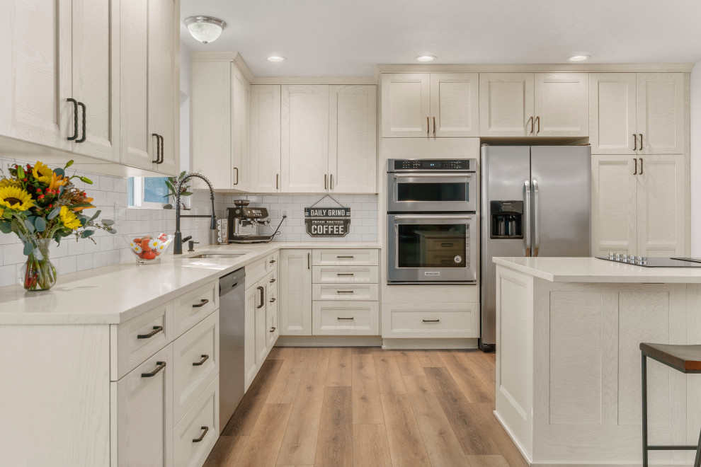 Photo of a medium sized contemporary kitchen/diner in Seattle with a double-bowl sink, shaker cabinets, white cabinets, engineered stone countertops, white splashback, porcelain splashback, stainless steel appliances, laminate floors, a breakfast bar, brown floors and white worktops.