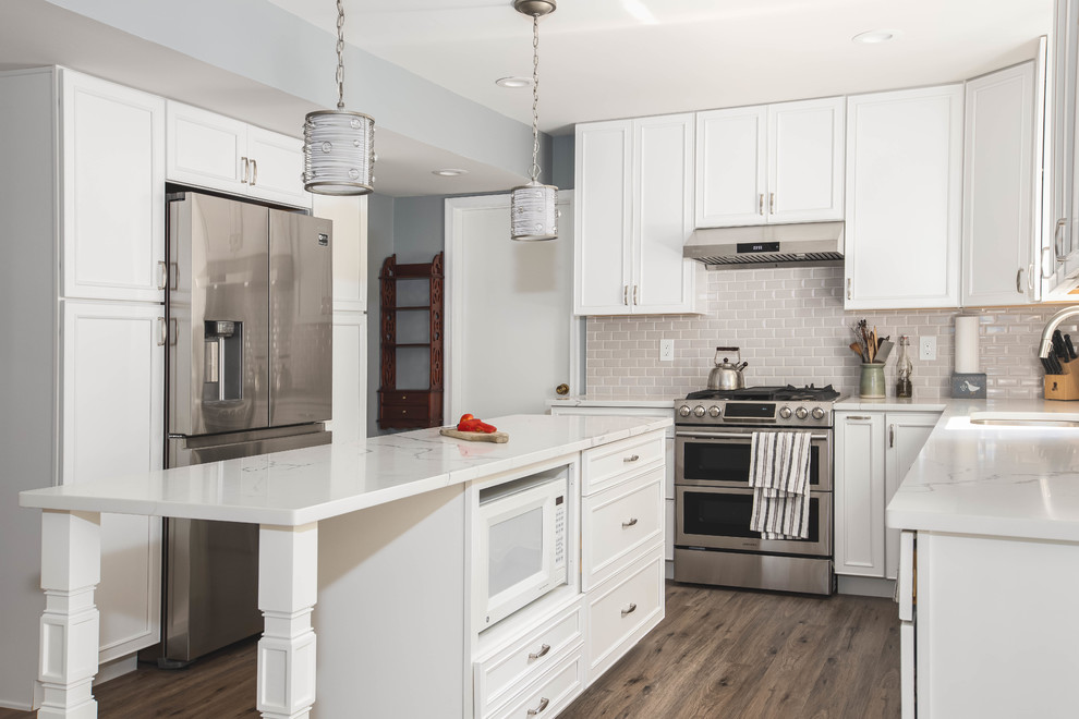 This is an example of a large contemporary u-shaped open plan kitchen in DC Metro with an undermount sink, raised-panel cabinets, white cabinets, quartz benchtops, grey splashback, subway tile splashback, stainless steel appliances, vinyl floors, with island, brown floor and white benchtop.