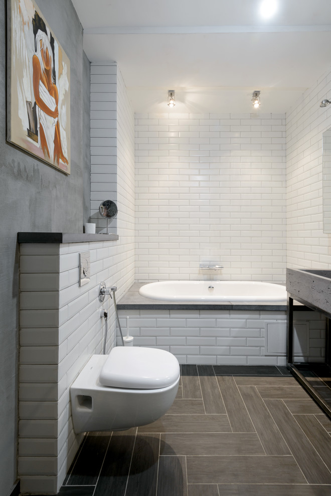 This is an example of a large industrial bathroom in Novosibirsk with a wall-mount toilet, white tile, grey walls, porcelain floors, a drop-in tub and subway tile.