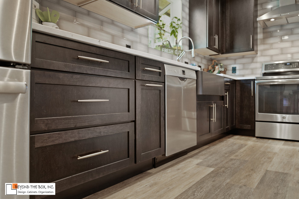 Mid-sized transitional l-shaped medium tone wood floor and brown floor kitchen photo with a farmhouse sink, shaker cabinets, black cabinets, granite countertops, gray backsplash, ceramic backsplash, stainless steel appliances, an island and white countertops