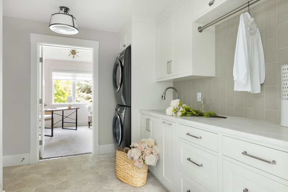 Photo of a small beach style galley dedicated laundry room in Minneapolis with a double-bowl sink, shaker cabinets, white cabinets, quartz benchtops, grey splashback, subway tile splashback, grey walls, porcelain floors, a stacked washer and dryer, grey floor and white benchtop.