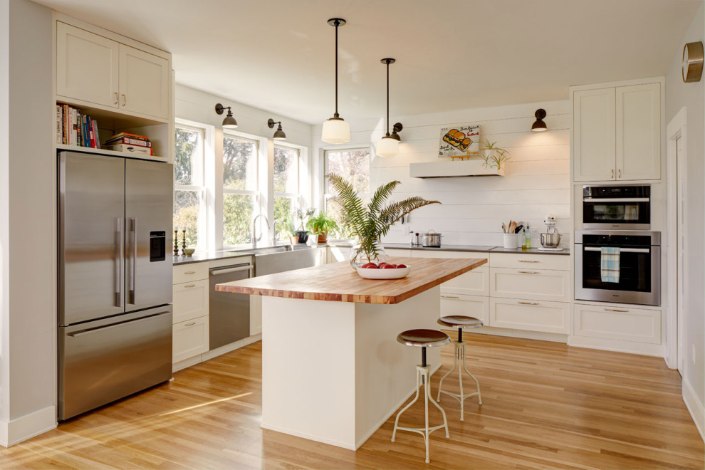 Photo of a country u-shaped kitchen in Seattle with a farmhouse sink, shaker cabinets, white splashback, stainless steel appliances, medium hardwood floors, brown floor and black benchtop.
