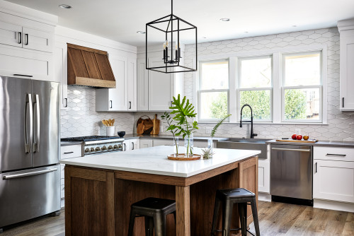 Contemporary farmhouse kitchen with white shaker RTA cabinets, wood range hood, stained-wood island with quartz top, hex-tile backsplash, black hardware and faucet, stainless appliances, and a lantern pendant.