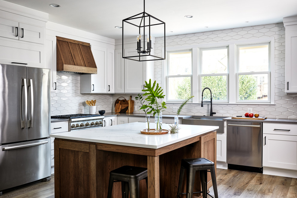 Diseño de cocina tradicional renovada de tamaño medio con electrodomésticos de acero inoxidable, suelo de madera en tonos medios, una isla, fregadero sobremueble, armarios estilo shaker, puertas de armario blancas, encimera de cuarzo compacto, salpicadero blanco, suelo marrón y encimeras grises