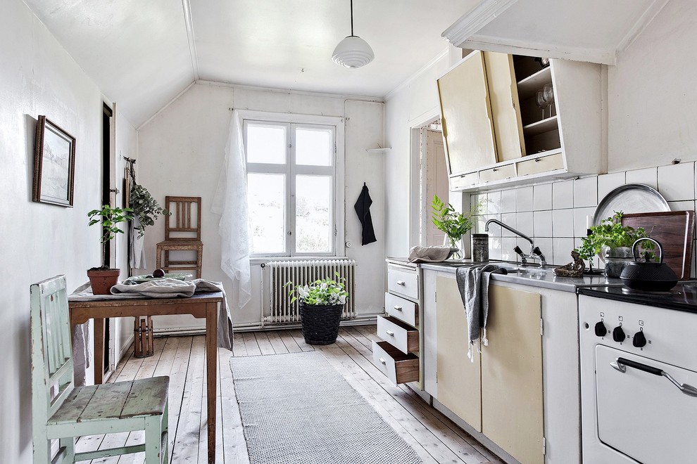 This is an example of a scandinavian single-wall eat-in kitchen in Gothenburg with an integrated sink, flat-panel cabinets, white cabinets, stainless steel benchtops, white splashback, white appliances, light hardwood floors, beige floor and grey benchtop.