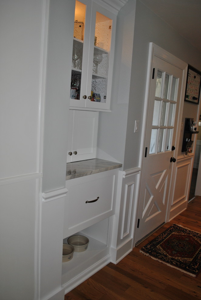 Example of a mid-sized transitional l-shaped medium tone wood floor eat-in kitchen design with a farmhouse sink, shaker cabinets, white cabinets, granite countertops, white backsplash, ceramic backsplash, stainless steel appliances and an island