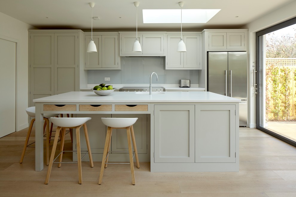Inspiration for a mid-sized transitional single-wall open plan kitchen in Surrey with an integrated sink, shaker cabinets, grey cabinets, quartz benchtops, white splashback, glass sheet splashback, stainless steel appliances, light hardwood floors, with island and beige floor.