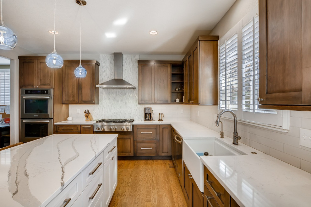 Photo of a large transitional u-shaped eat-in kitchen in Denver with a farmhouse sink, shaker cabinets, brown cabinets, quartz benchtops, white splashback, stainless steel appliances, medium hardwood floors, with island, brown floor and white benchtop.
